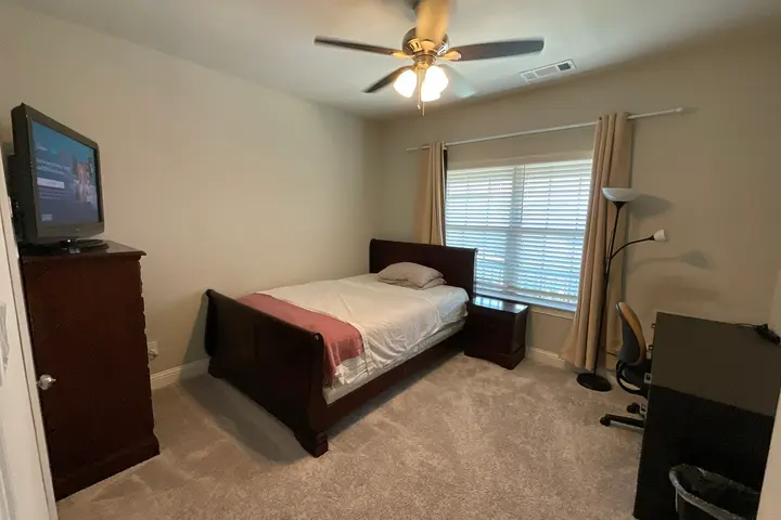 Queen bed by the curtained window with a TV on the dresser and a desk